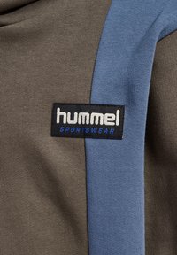 Hummel-Sportbekleidungsetikett auf einem Stoff mit braunen und blauen Abschnitten, der eine strukturierte Oberfläche aufweist, und einem rechteckigen schwarzen Aufnäher mit weißem und blauem Text.