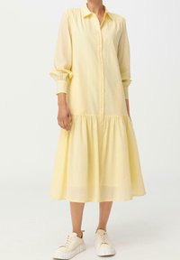 Żółta sukienka zapinana na guziki, z długimi rękawami, kołnierzykiem koszulowym i marszczonym pasem. Spódnica jest rozkloszowana, wykonana z lekkiego materiału. Stylizowana z białymi trampkami.