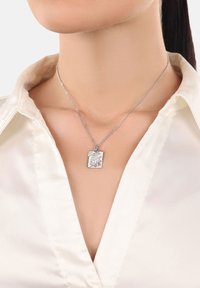 Collana con ciondolo in argento su una catena sottile, caratterizzata da un design inciso e forma rettangolare, indossata su una camicetta di satin bianca.