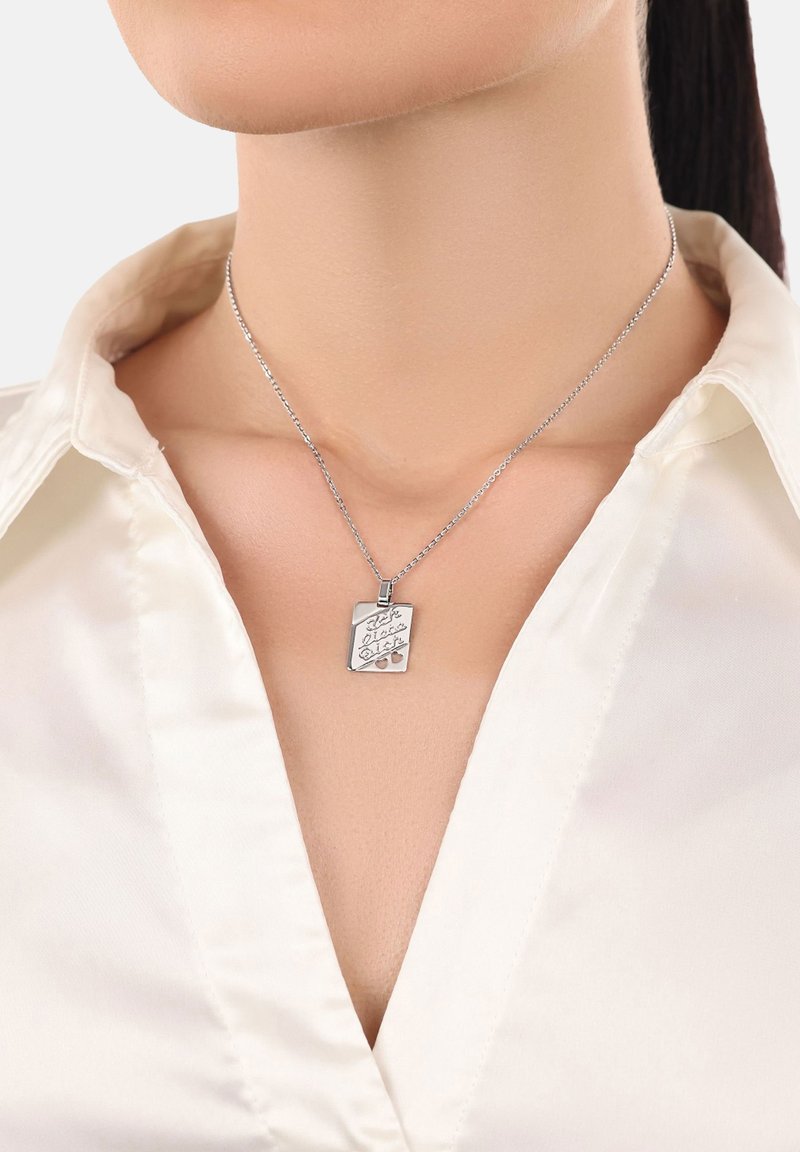 Collana con ciondolo in argento su una catena sottile, caratterizzata da un design inciso e forma rettangolare, indossata su una camicetta di satin bianca.