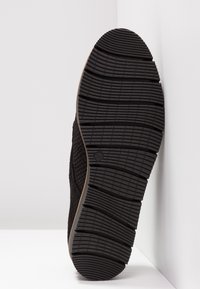 Semelle d'une chaussure noire avec un motif de bande de roulement texturé, positionnée debout contre un coin blanc mur et sol.