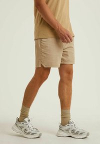 Beige šortky vyrobené z lehké texturované tkaniny; mají šikmé postranní kapsy a mírně zakřivený lem. Doplňují je šedé tenisky a béžové ponožky.