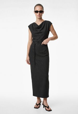 Kvinde iført en ærmeløs mørk pinstripet midi-kjole med et cowl-halsudskæring, tyndt bælte, sorte hælede sandaler, solbriller og pandebånd, stående.