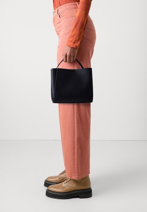 Personne portant un pantalon rose et des chaussures beiges à lacets, tenant un sac à main noir par sa poignée supérieure sur un fond uni.