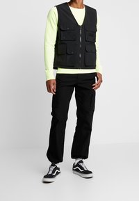 Svart verktygsväst med flera fickor, bärs över en neongrön långärmad tröja, i kombination med svarta cargobyxor och svarta sneakers.