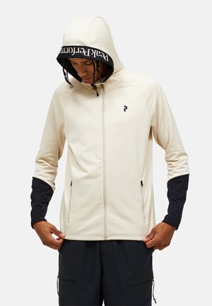 Persona che indossa una felpa con cappuccio beige con polsini neri e fodera del cappuccio, mentre sistema l'orlo inferiore della felpa guardando verso il basso.