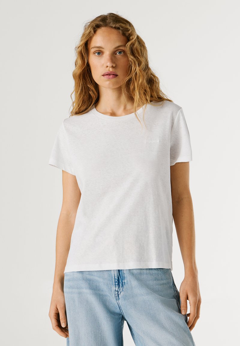 Kvinde med bølget blondt hår iført en ensfarvet hvid kortærmet T-shirt og lyseblå jeans mod en neutral baggrund.