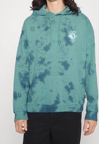 Sudadera con capucha color verde azulado con patrón de tie-dye y bolsillo frontal. Presenta un logo en el pecho y puños y dobladillo acanalados. Textura de tela suave.