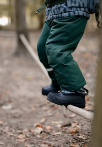 Botas impermeables negras con suelas texturizadas y pantalones impermeables verdes. Niño equilibrándose sobre un tronco en un entorno natural al aire libre.