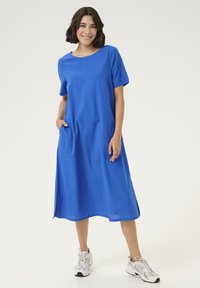 Blaues Leinenkleid mit kurzen Ärmeln, lockerer Passform, rundem Halsausschnitt, Seitentaschen und knielangem Saum. Getragen mit weißen Sneakers.