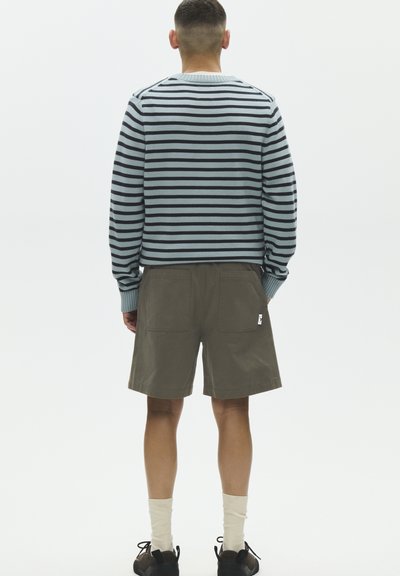 Homme portant une chemise à rayures à manches longues, un short vert olive, des chaussettes blanches et des chaussures marron, vu de dos sur un fond uni.