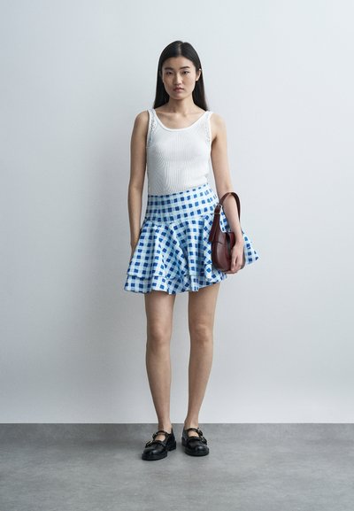 Γυναίκα που στέκεται μπροστά σε γκρι τοίχο, φορώντας λευκό τοπάκι, μπλε και λευκή καρό φούστα, μαύρα παπούτσια και κρατώντας καφέ τσάντα στον βραχίονά της.