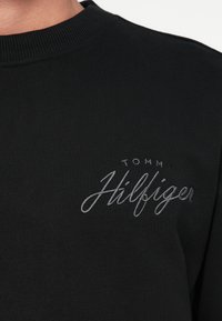 Črna majica s okroglim ovratnikom in sivi vezeni logotip "Tommy Hilfiger" na levem prsnem delu.
