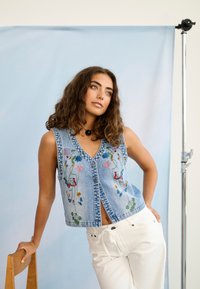 Femme aux cheveux bouclés portant un gilet en denim bleu brodé et un pantalon blanc, appuyée sur une chaise en bois devant un fond bleu clair.