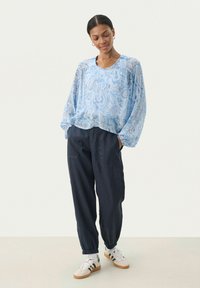 Blusa azzurra chiaro con motivo paisley e maniche lunghe, abbinata a pantaloni scuri a sigaretta blu navy e sneakers bianche con strisce nere.