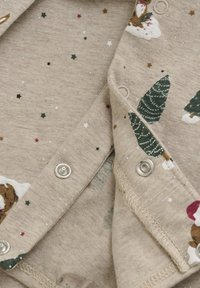 Tissu beige présentant un motif d'arbres de Noël, de flocons de neige et d'étoiles. Comprend des pressions métalliques le long de l'encolure. Texture douce.