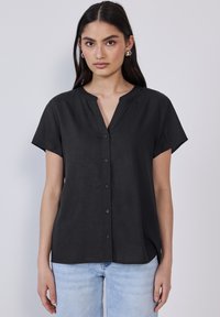 Sort kortærmet bluse med V-hals og frontknapper; glat tekstur og afslappet pasform. Bærers med lyseblå jeans.