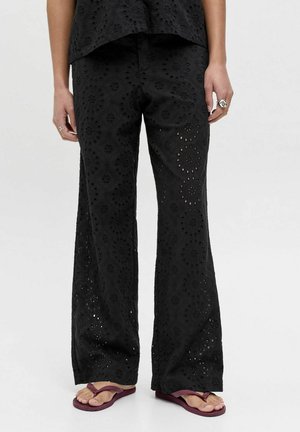 Kvinne iført svarte vide bukser med øyeletthull og sirkelmønstre, ringer på fingrene, og mørkerøde flip-flops, står foran en ensfarget bakgrunn.