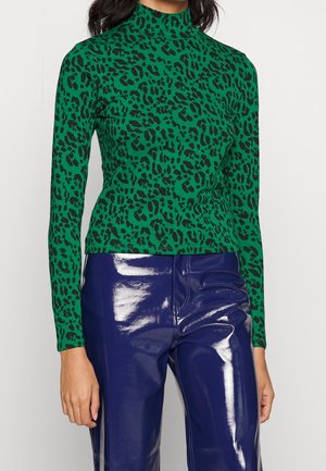 Femme portant un haut à manches longues imprimé léopard vert et un pantalon taille haute bleu marine brillant, debout devant un fond uni.