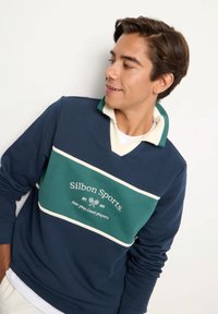 Giovane uomo che indossa un felpa sportiva Silbon blu navy e verde con logo del tennis, guarda verso destra su uno sfondo bianco semplice.