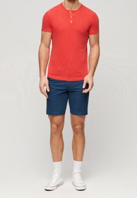 Chemise henley rouge en coton à manches courtes avec trois boutons, assortie à un short bleu en coton. Le mannequin porte des baskets blanches et des chaussettes montantes jusqu'à la cheville.