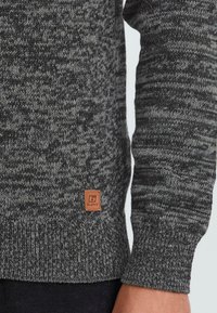 Primer plano de una persona que viste un suéter de punto gris y negro con una pequeña etiqueta marrón de la marca "Blend" cerca del dobladillo inferior.