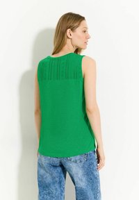 Top verde senza maniche con una sezione superiore testurizzata e stampata; tessuto morbido, vestibilità ampia. Abbinato a jeans di denim blu chiaro.