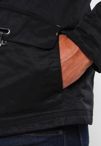 Sort, let jakke med en glat tekstur, der har en lynlås-lomme og synlige syninger. Båret over denim jeans.