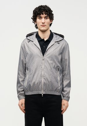 Jeune homme aux cheveux bouclés portant une veste à capuche zippée argentée brillante par-dessus une chemise noire à col, debout devant un fond uni.