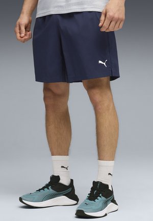 Hombre con pantalones cortos azul marino de Puma, calcetines blancos de Puma y zapatillas azul y negro de Puma, de pie sobre un fondo gris.