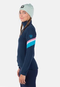 Námořnický fleecový pulovr s vysokým límcem, se vzorem modré, růžové a bílé proužky na rukávu a logem na rameni.