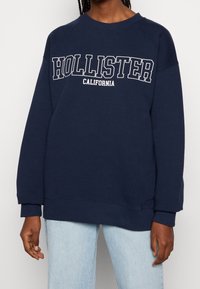 Granatowa bluza z bawełny, z okrągłym dekoltem i ściągaczami na mankietach. Na przodzie znajduje się napis "HOLLISTER CALIFORNIA" w białych literach.