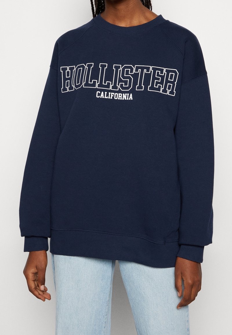 Granatowa bluza z bawełny, z okrągłym dekoltem i ściągaczami na mankietach. Na przodzie znajduje się napis "HOLLISTER CALIFORNIA" w białych literach.