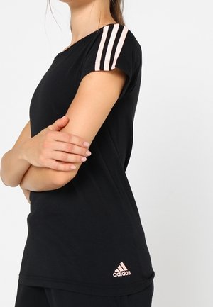 Frau mit schwarzem Adidas-T-Shirt mit pinkfarbenen Streifen an den Ärmeln und pinkfarbenem Logo, verschränkte Arme, vor einfarbigem weißem Hintergrund.