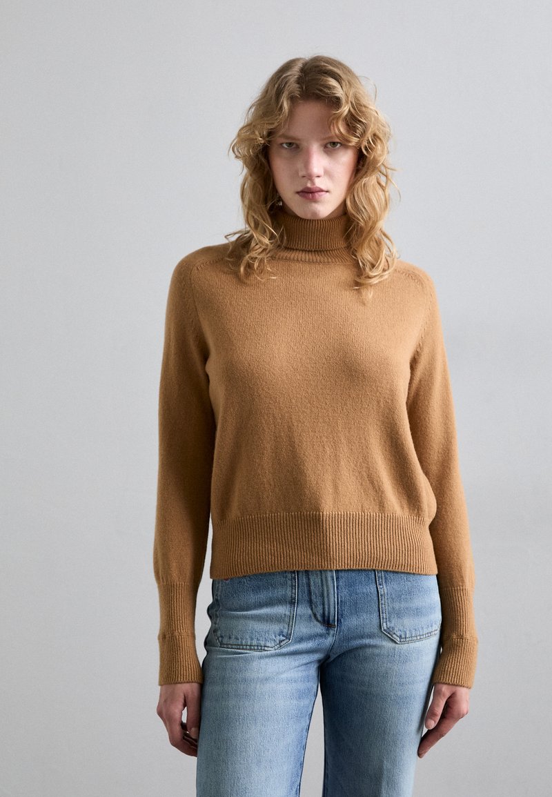 Teve színű pulóver magas gallérral, bordás mandzsettákkal és aljjal, világoskék farmernadrággal párosítva. Az anyag lágy és textúrált hatású.