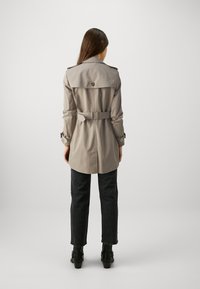 Abrigo trench beige con diseño de doble botonadura, cintura con cinturón, hombreras y puños de manga enrollados, combinado con vaqueros negros y botas de tacón.