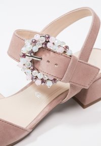 Sandale en daim rose avec un talon épais, présentant une boucle décorative en perles dans des teintes bleu clair, rose et violet. Détail intérieur lisse visible.