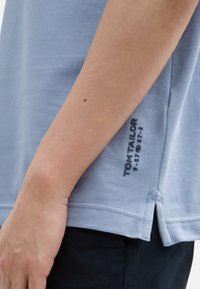 Svetlomodré tričko s krátkymi rukávmi vyrobené z bavlny. Obsahuje bočný rozparok na okraji a tmavomodrý potlačený logotyp značky na spodnej strane.