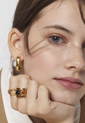 Pendiente de aro dorado, dos anillos dorados con detalles texturizados y una gema negra. Textura de piel suave, color de labios rosa y maquillaje suave.