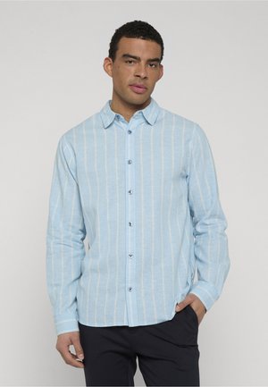 Mann trägt ein hellblaues Hemd mit langen Ärmeln und vertikalen weißen Streifen sowie eine dunkle Hose, eine Hand in der Tasche, neutraler Hintergrund.