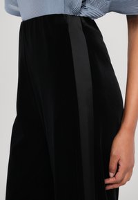 Pantalons en velours noir avec une rayure noire brillante sur le côté. Le tissu semble doux, avec une coupe ample et un haut bleu en maille partiellement visible.