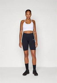 Sujetador deportivo blanco combinado con pantalones cortos de bicicleta negros de talle alto. La modelo lleva calcetines y zapatos negros, mostrando un look atlético ceñido al cuerpo.