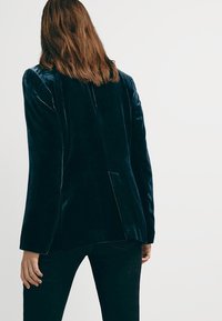 Femme aux cheveux bruns mi-longs portant un blazer en velours bleu marine et un pantalon noir, vue de dos sur un fond uni clair.
