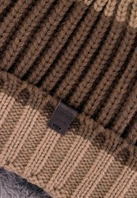 Strickmütze in Braun und Beige, mit gerippter Textur, umgeschlagenem Saum und einem kleinen schwarzen Leder-Logosticker an der Seite.