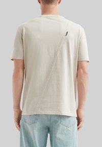 Jasno beżowy t-shirt z bawełny z unikalnym ukośnym szwem na plecach i krótkimi rękawami, w parze z jasno niebieskimi dżinsami.