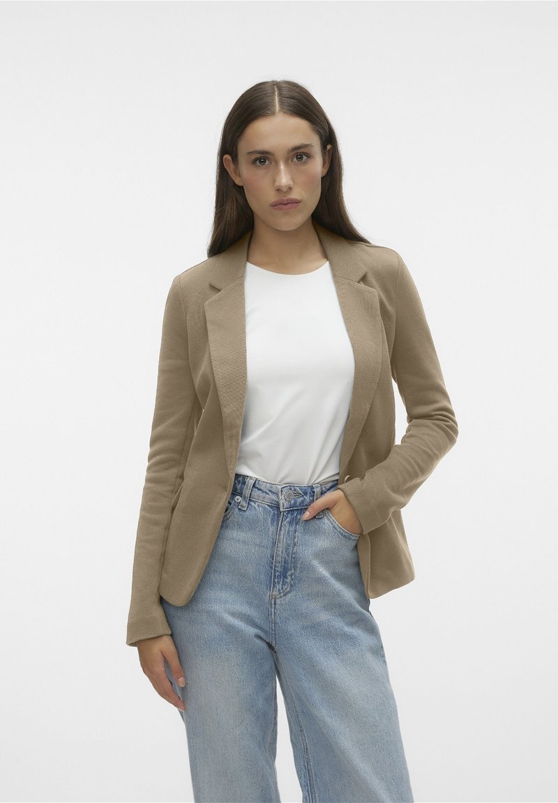 Blazer beige ajustado hecho de tela suave, con solapas muescas y mangas largas, combinado con una camisa blanca y jeans de color azul claro.