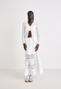 Woman with braided hair wearing a white buttoned shirt, long eyelet skirt, silver sneakers, and hoop earrings against a light background.