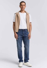 Maglione beige senza maniche sopra una t-shirt bianca, abbinato a jeans blu scuro e sneakers bianche; posa in piedi, sfondo neutro.