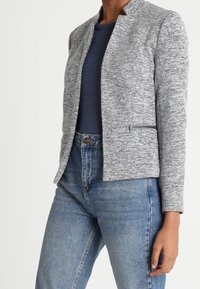 Šedý texturovaný blazer s vystouplým límcem a předními kapsami na zip, nošený přes námořnický úzký top, kombinovaný s vysokými modrými džínami.