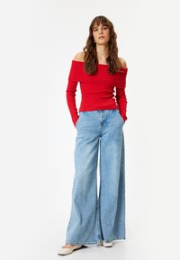Maglione rosso a spalle scoperte con maniche lunghe abbinato a jeans in denim azzurri a gamba larga. I jeans hanno un design classico a cinque tasche.